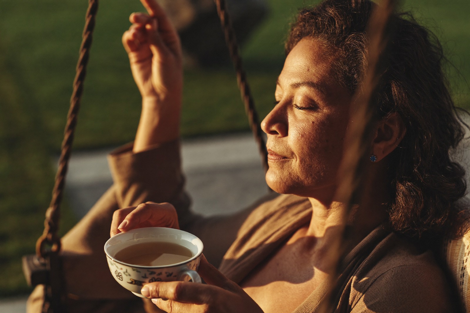 A woman rests with her eyes closed on a porch swing, holding a cup of tea
