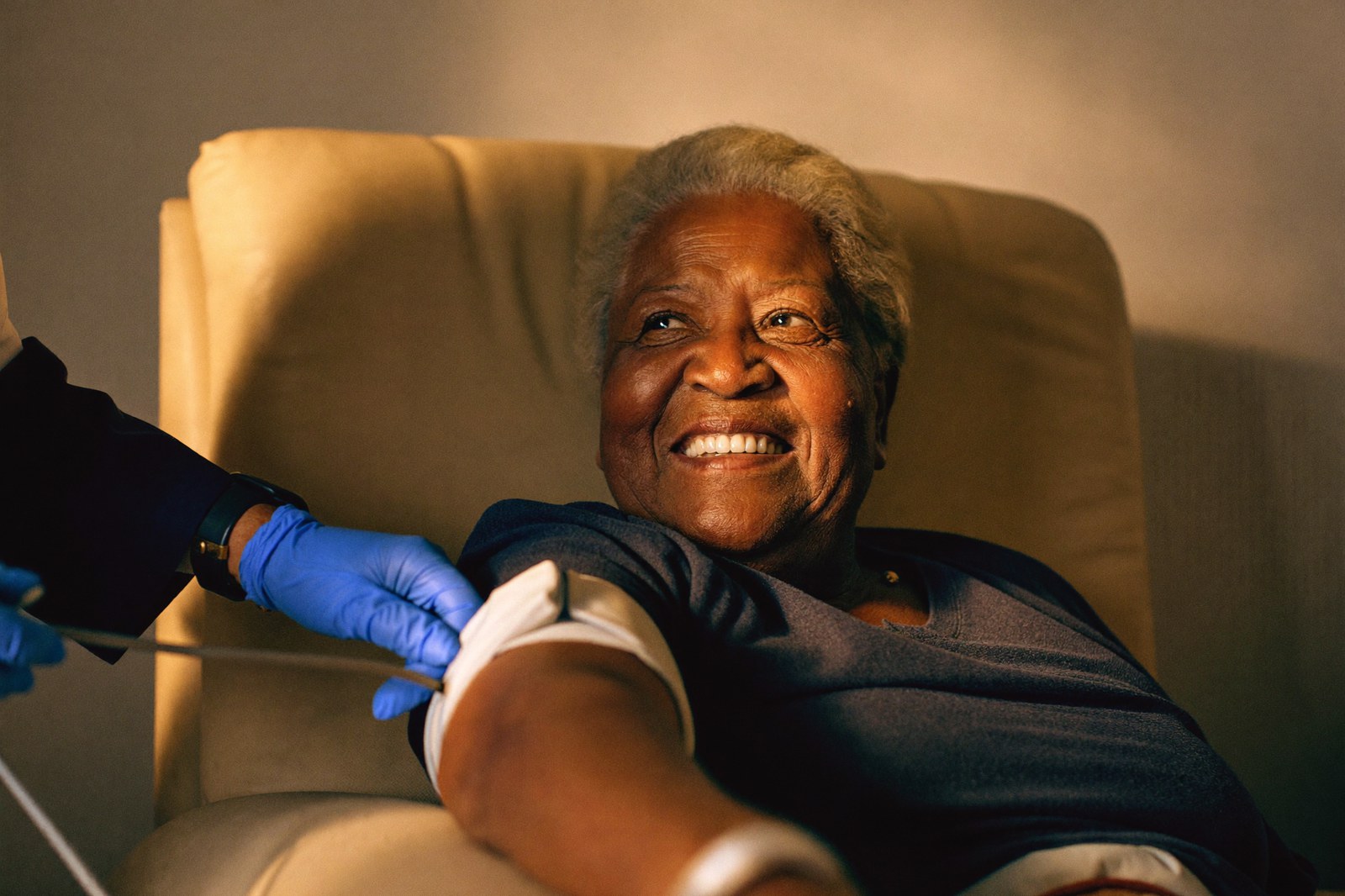 A smiling woman sits in an infusion recliner as a nurse fits a blood pressure cuff